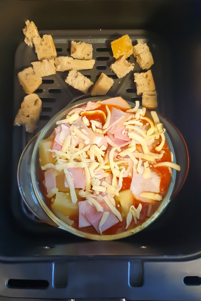 A glass dish brimming with ham, cheese, and pineapple transforms into a delectable Pizza Bowl in the air fryer, surrounded by small pieces of bread.