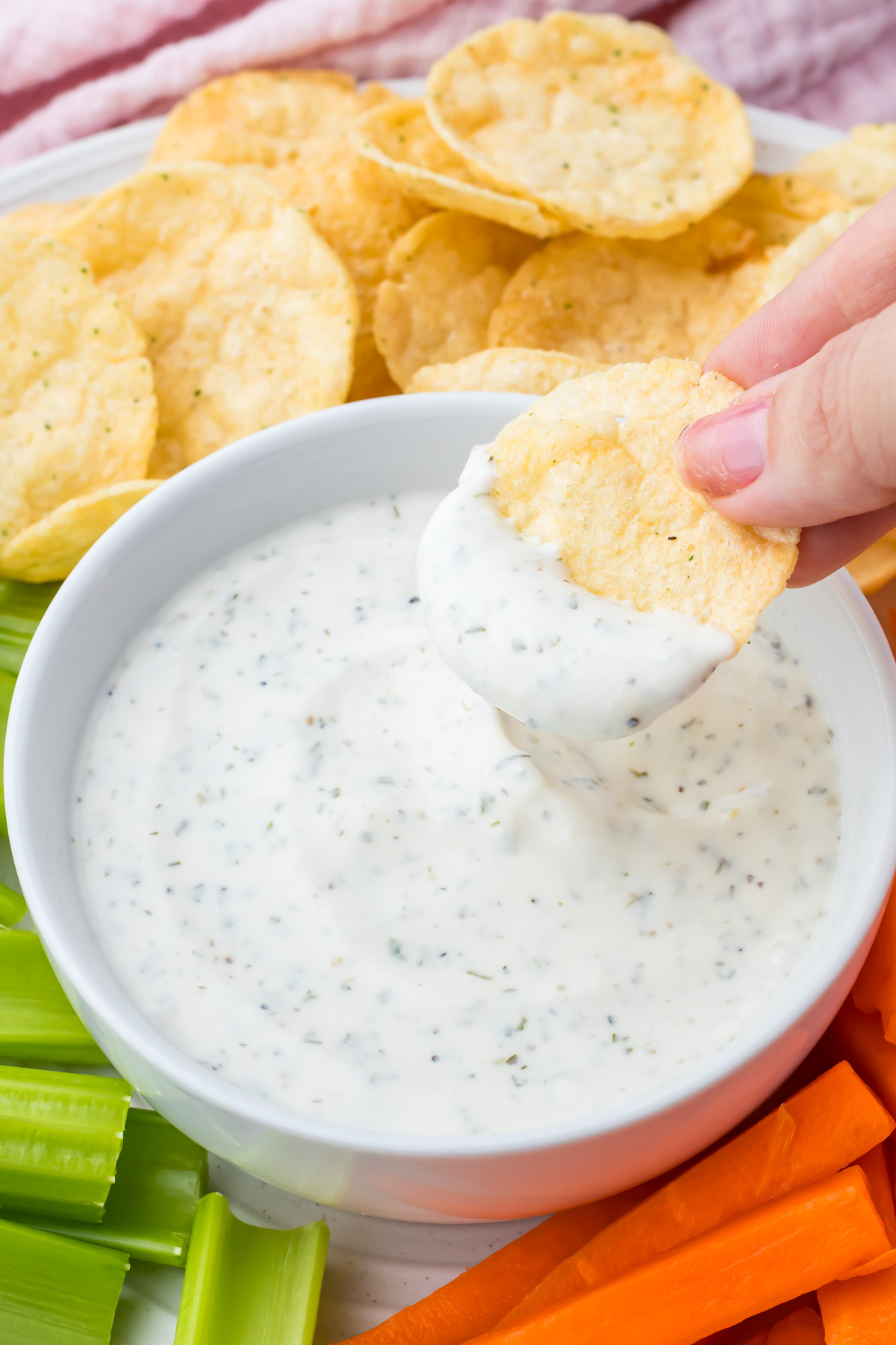 A hand dips a round chip into a creamy cottage cheese ranch dip, surrounded by fresh celery and carrot sticks.
