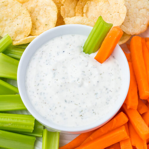 A bowl of creamy ranch dip is surrounded by crisp celery sticks, carrot sticks, and round chips.