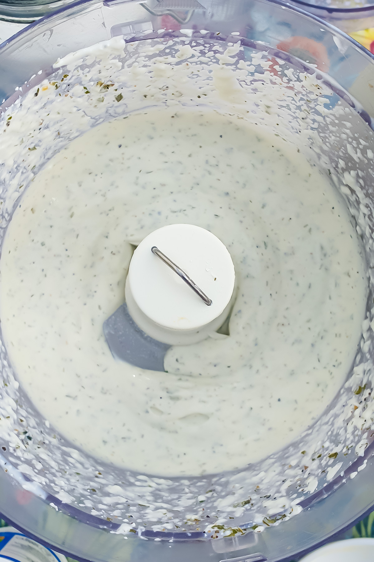 Close-up of a food processor bowl containing a creamy, white mixture with specks of green herbs, likely a ranch dip or dressing made with cottage cheese, around a central blade mechanism.