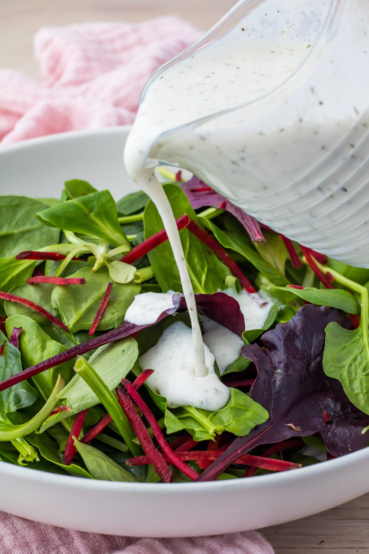 A refreshing mixed green salad with spinach, beet strips, and leafy greens is elegantly drizzled with creamy cottage cheese ranch dressing in a white bowl.