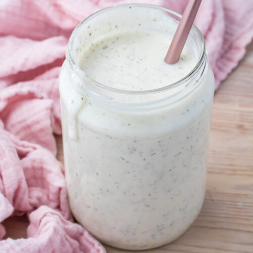 A jar of cottage cheese ranch dressing blended with cottage cheese and visible herbs sits on a wooden surface, next to a pink cloth. A stainless steel spoon is placed in the jar.