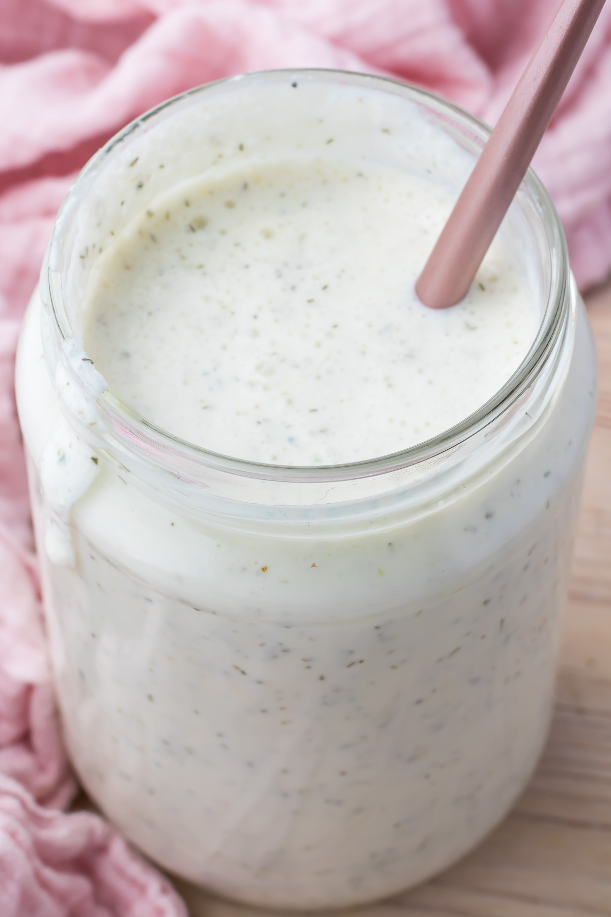 A jar of creamy cottage cheese ranch dressing with herbs and a spoon rests on a wooden surface, complemented by a soft pink cloth.