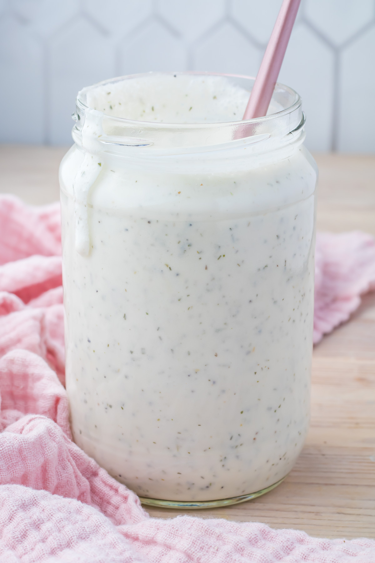A glass jar filled with creamy, speckled cottage cheese ranch dressing, with a pink spoon inside. The jar rests on a wooden surface, accompanied by a pink cloth.