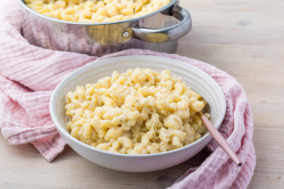 A bowl of creamy high-protein macaroni and cheese sits on the table, complemented by a pink cloth and a pot in the background.