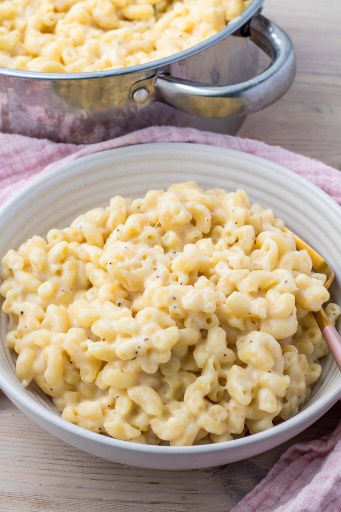 A white bowl filled with creamy, high-protein macaroni and cheese sits on a wooden surface. A pot of the same dish is partially visible in the background.