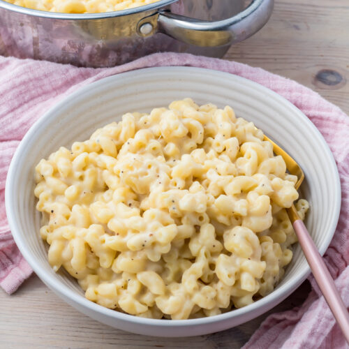 A bowl of creamy, high-protein macaroni and cheese with a fork rests on a pink cloth next to a pot on a wooden surface.