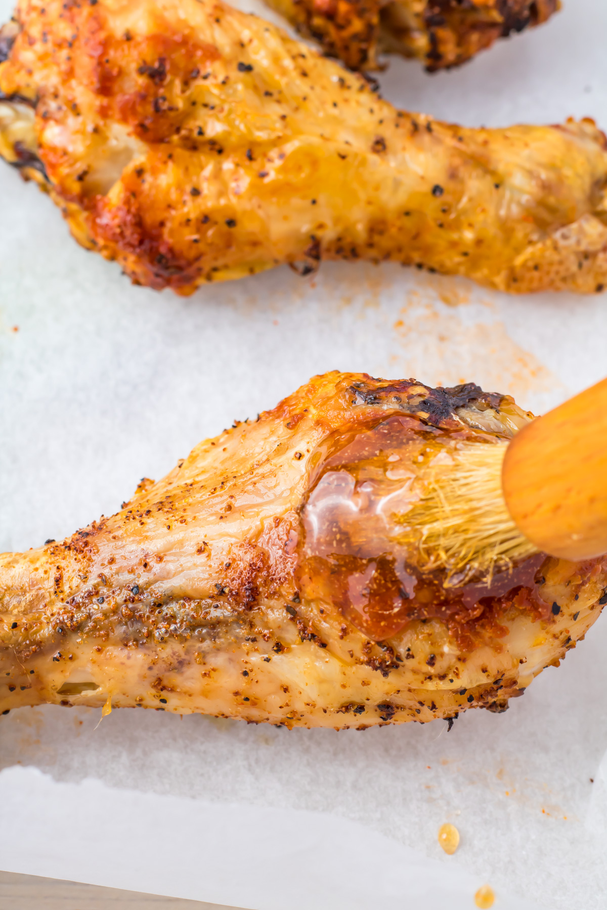 Close-up of a Hot Honey Chicken drumstick being brushed with sauce on parchment paper, seasoned with pepper and spices.