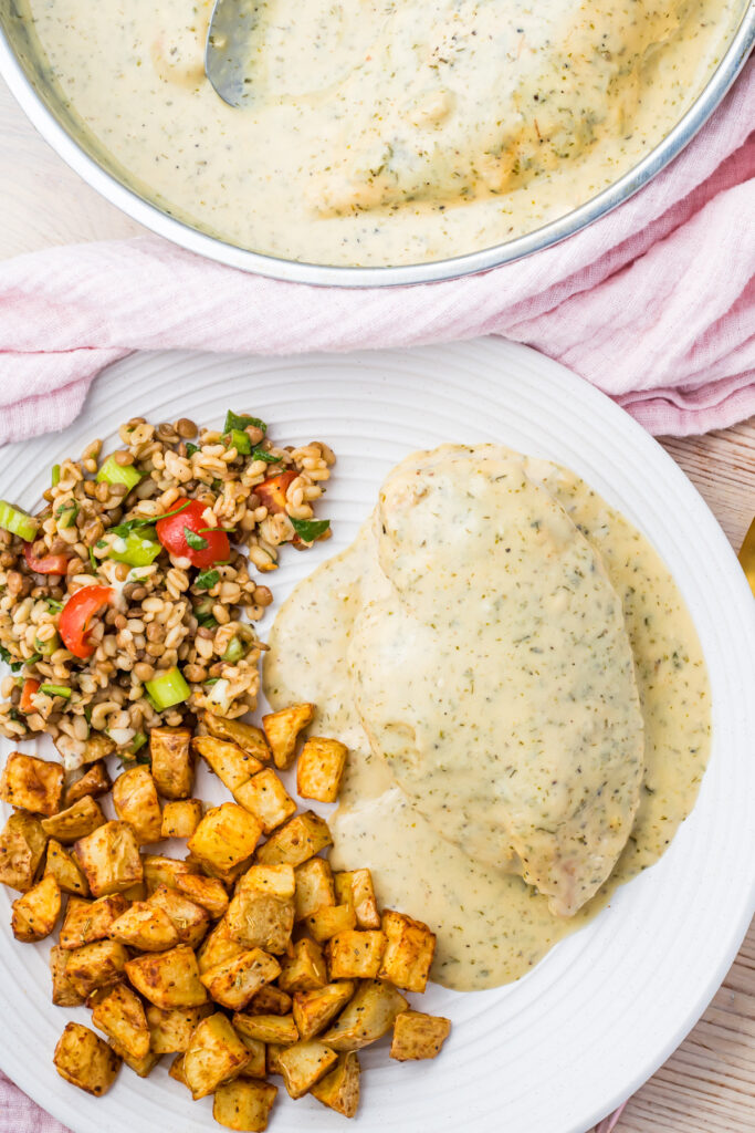 A plate showcases Ranch chicken covered in creamy sauce, accompanied by diced roasted potatoes and a mixed grain salad with tomatoes and herbs. Nearby, a pan holds extra sauce and chicken, resting on a pink cloth.
