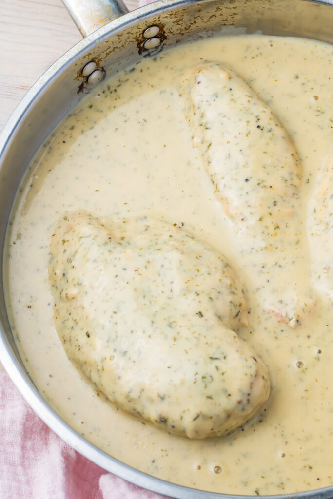 Chicken breasts simmering in a creamy herb and ranch sauce in a metal pan.
