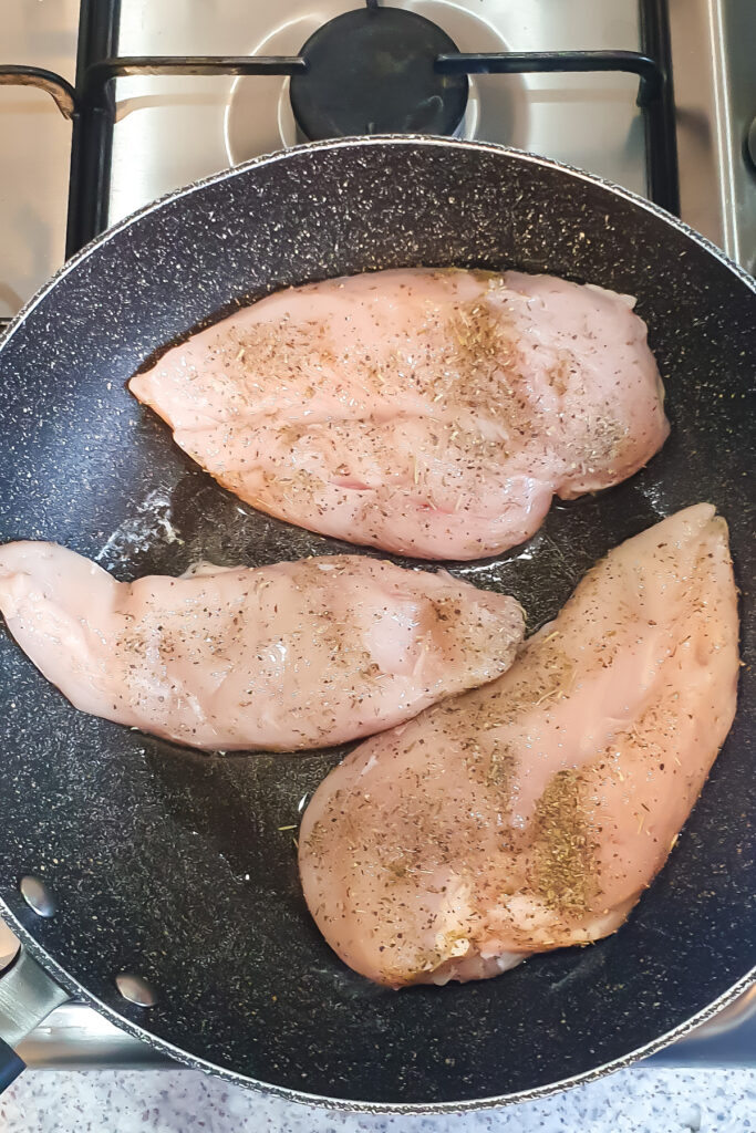 Three raw ranch chicken breasts, seasoned with herbs, sizzle in a black frying pan on a gas stove.