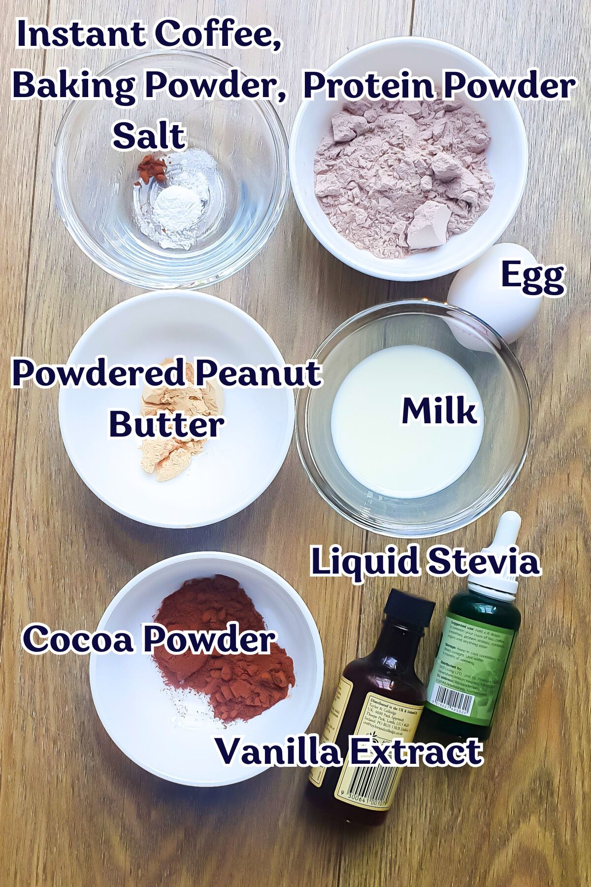Assorted baking ingredients for a chocolate protein mug cake in bowls and bottles on a wooden surface, each labeled: instant coffee, baking powder, protein powder, salt, egg, powdered peanut butter, milk, liquid stevia, cocoa powder, butter, vanilla extract.