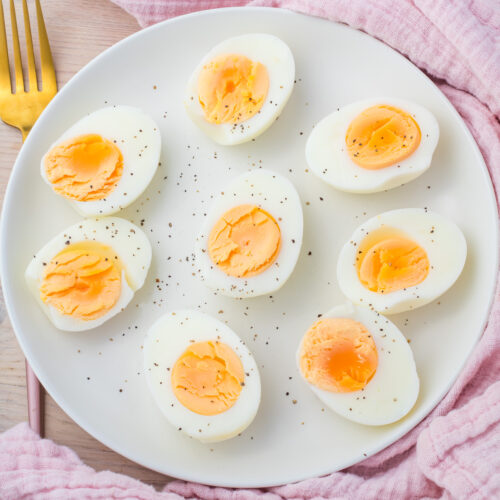 A white plate with nine halved Air Fryer Boiled Eggs sprinkled with black pepper sits on a pink cloth, with a gold fork beside it.