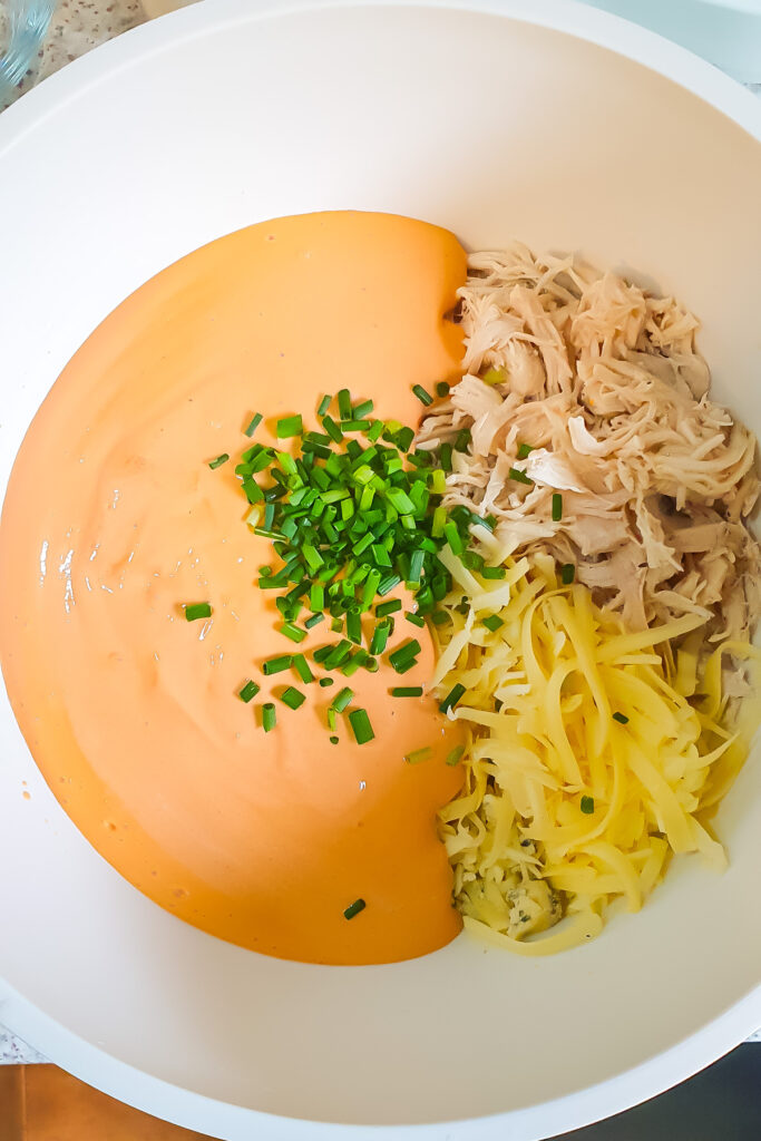 A white bowl containing shredded chicken, shredded cheese, buffalo cottage cheese sauce, and chopped chives arranged in separate sections.