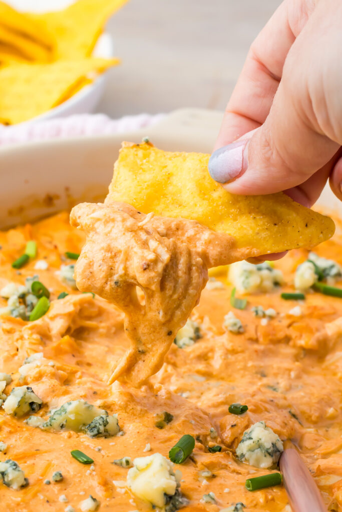 A hand dips a tortilla chip into a creamy Buffalo Chicken Dip with cottage cheese and topped with blue cheese crumbles and chopped green onions.