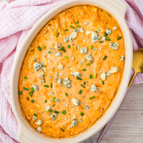 An oval baking dish filled with creamy Buffalo Chicken Dip with cottage cheese, topped with blue cheese crumbles and chopped chives, sits on a pink cloth next to a gold spoon.
