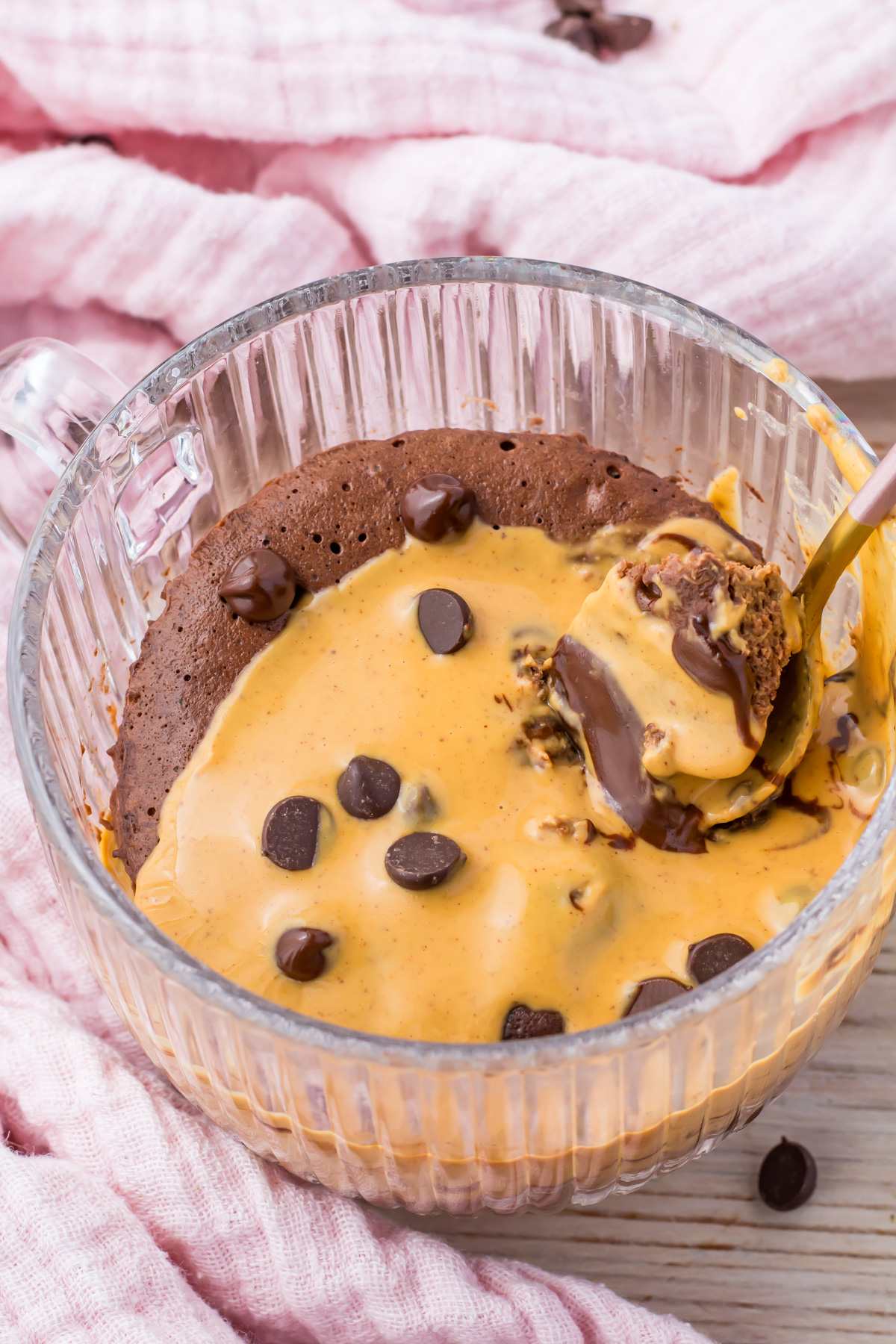 A glass mug filled with a rich Chocolate Protein Mug Cake topped with melted peanut butter and chocolate chips, with a spoon scooping out a warm, delicious bite.