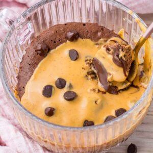 A glass bowl containing a Chocolate Protein Mug Cake topped with melted peanut butter and chocolate chips, with a spoon scooping some out.