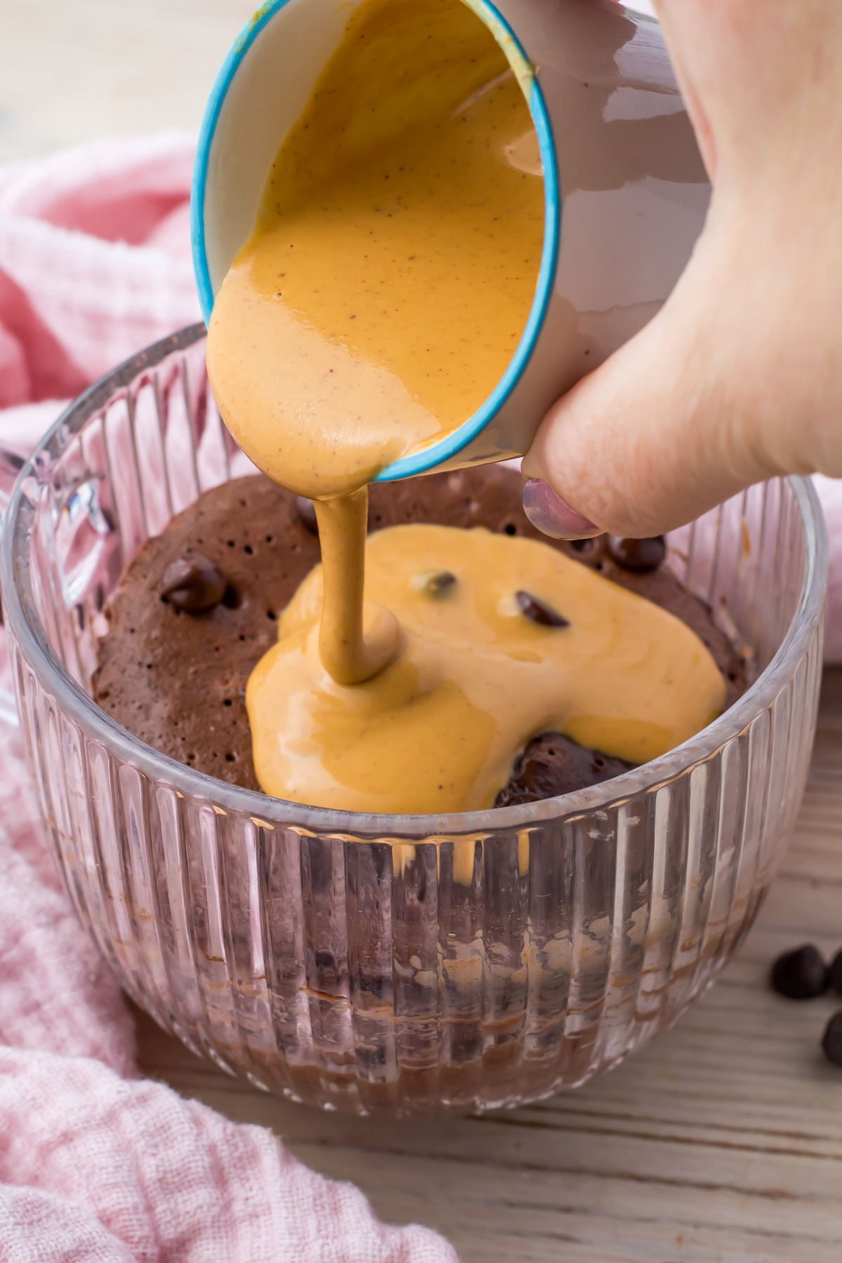 A hand pours creamy peanut butter sauce from a cup onto a chocolate protein mug cake with chocolate chips in a glass bowl.