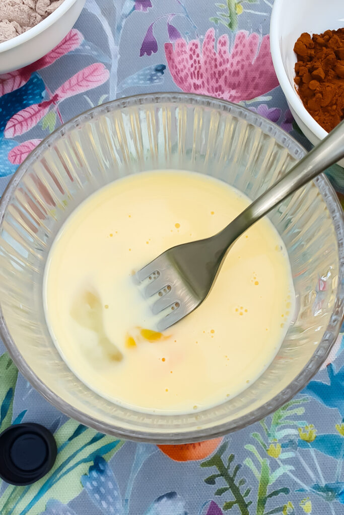 A metal fork rests in a glass bowl containing a mixture of beaten eggs and milk, perfect for preparing a chocolate protein mug cake, all placed on a floral-patterned tablecloth with other bowls nearby.