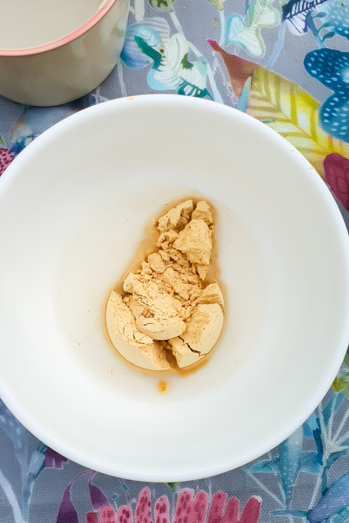 A white bowl containing a clump of light brown powdered peanut butter, partially dissolved in a small amount of liquid, is placed on a colorful floral-patterned surface.