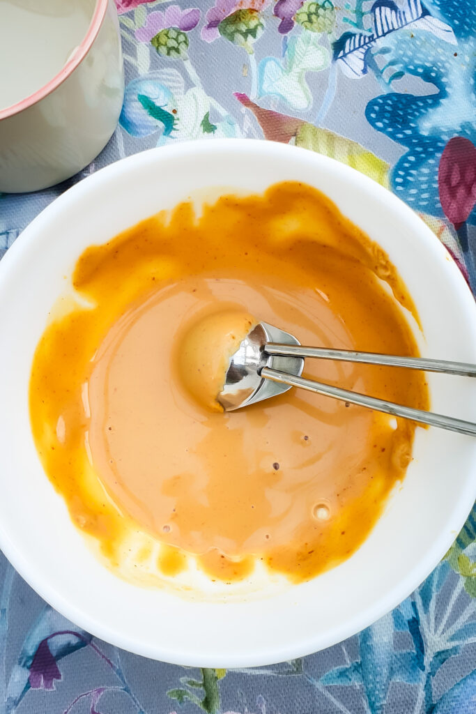 A metal scoop rests in a white bowl containing peanut sauce, set on a floral-patterned tablecloth.