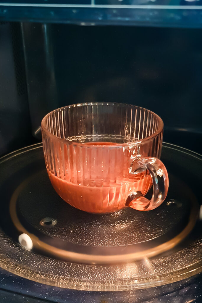 A glass cup filled with chocolate protein mug cake mixture sits on the turntable inside a microwave oven.