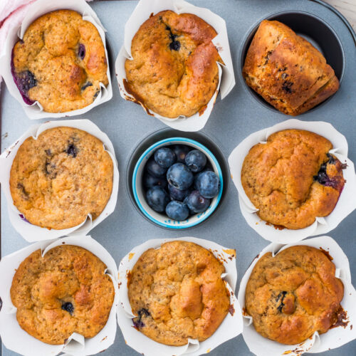 A tray with eight cottage cheese blueberry muffins in white paper liners and a small bowl of fresh blueberries in the center.