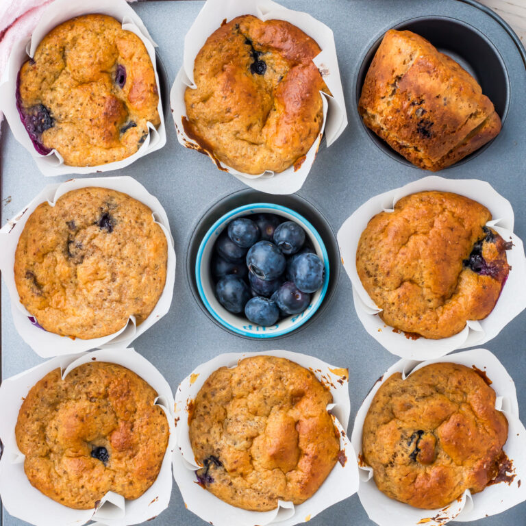 A tray with eight cottage cheese blueberry muffins in white paper liners and a small bowl of fresh blueberries in the center.