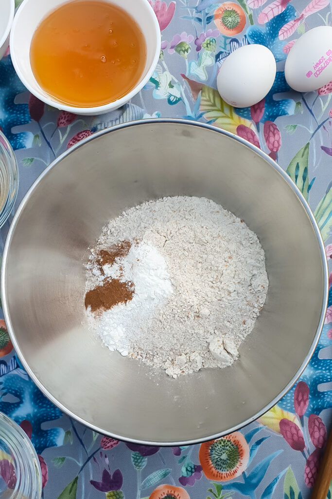 A metal mixing bowl with flour, baking powder, and cinnamon, sits beside honey and eggs.