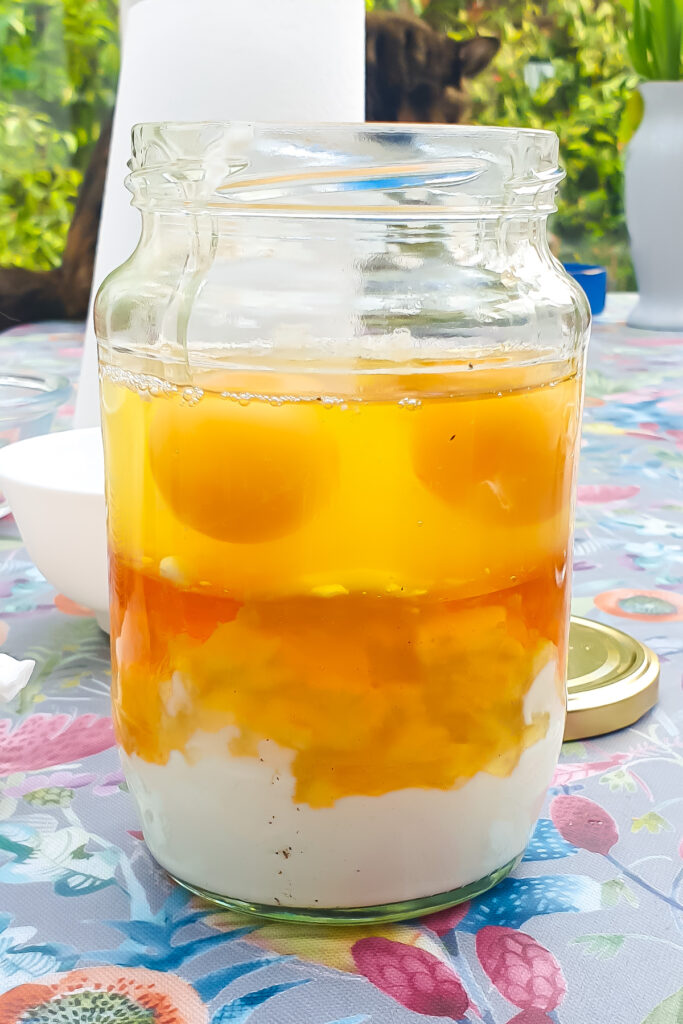 A glass jar containing unbroken raw eggs, including yolks and whites, and other wet ingredients, sits on a patterned tablecloth outdoors with the lid beside it.