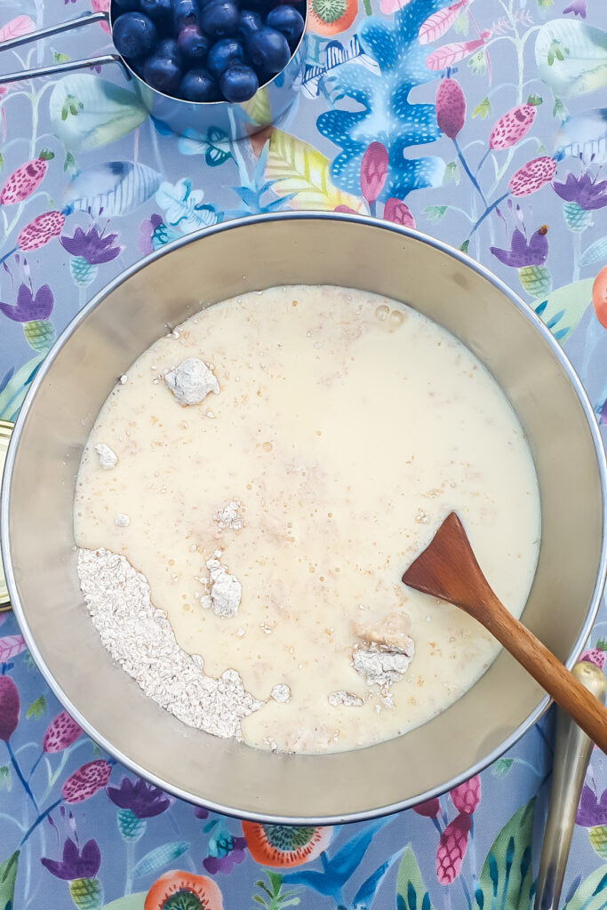 A mixing bowl with ingredients and a wooden spoon on a colorful tablecloth; a bowl of blueberries.