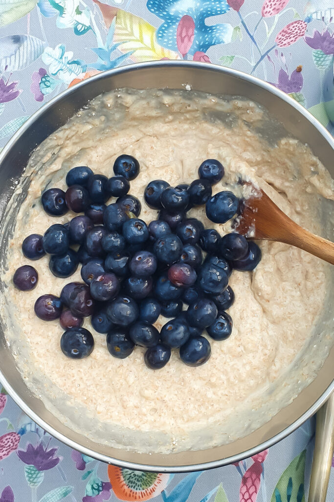 A mixing bowl with batter for cottage cheese Blueberry Muffins and a pile of fresh blueberries on top, being stirred with a wooden spoon, on a floral-patterned tablecloth.