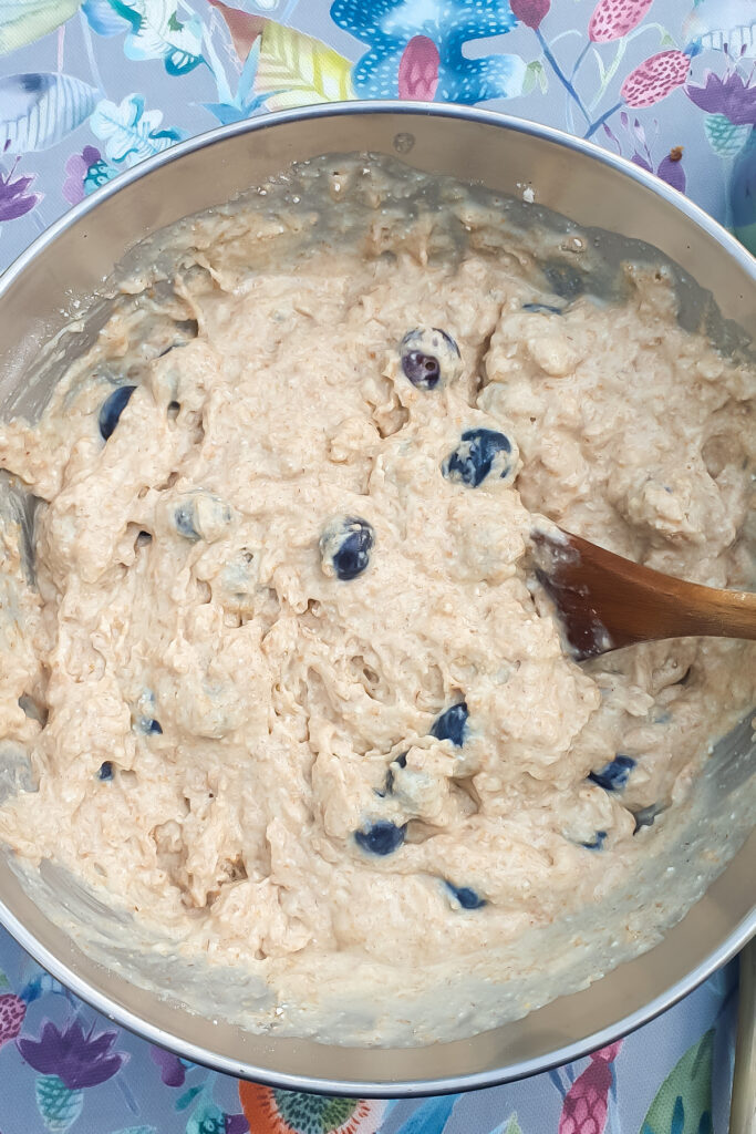 Bowl of thick, unbaked batter for Cottage Cheese Blueberry Muffins with visible blueberries being mixed with a wooden spoon on a floral-patterned surface.