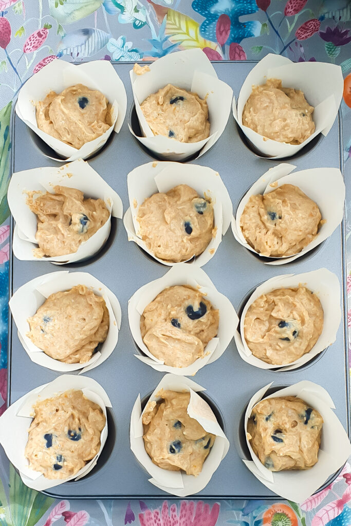 A muffin tray holds twelve paper liners, each filled with unbaked blueberry muffin batter with visible blueberries, resting on a colorful floral-patterned surface.