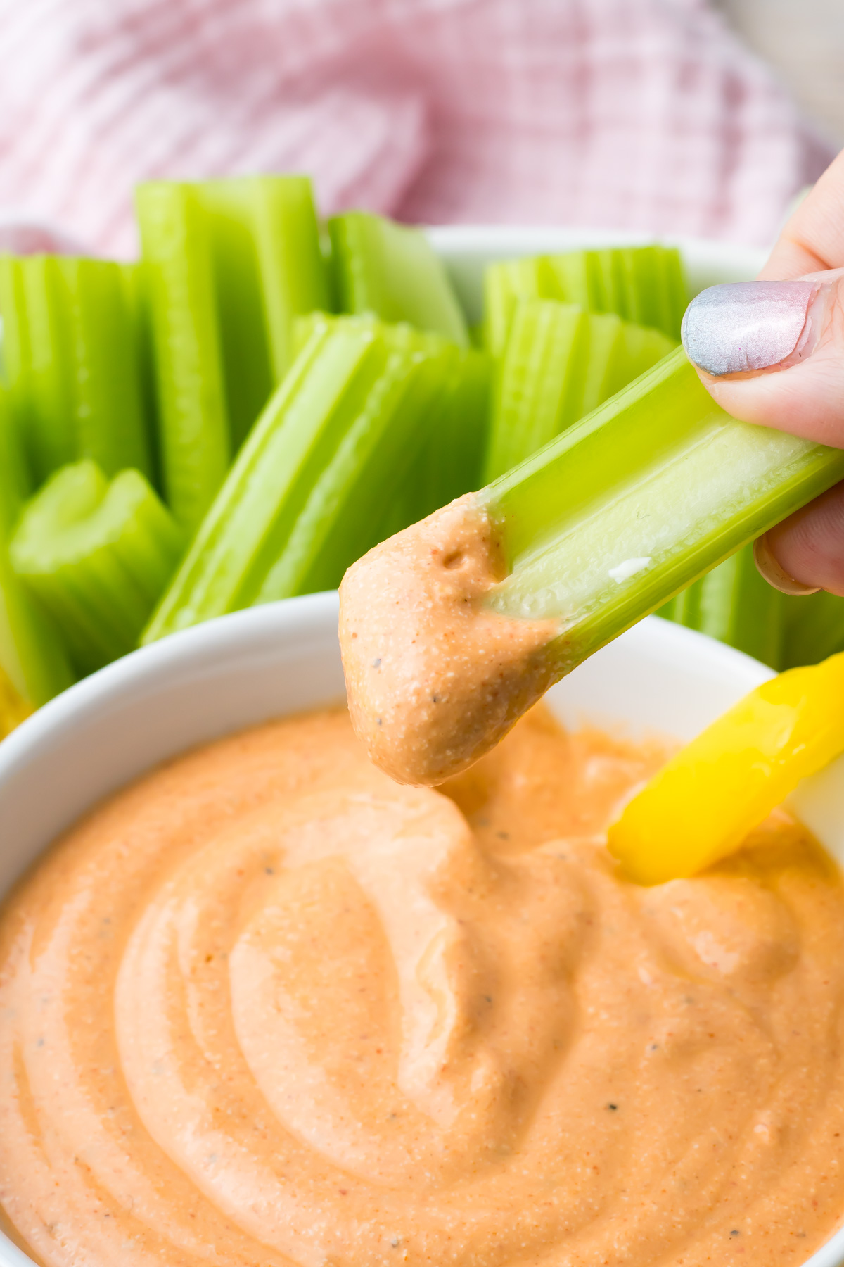 A hand dips a celery stick into a bowl of creamy orange Buffalo Dip made with cottage cheese, with more celery sticks and a yellow pepper slice in the background.