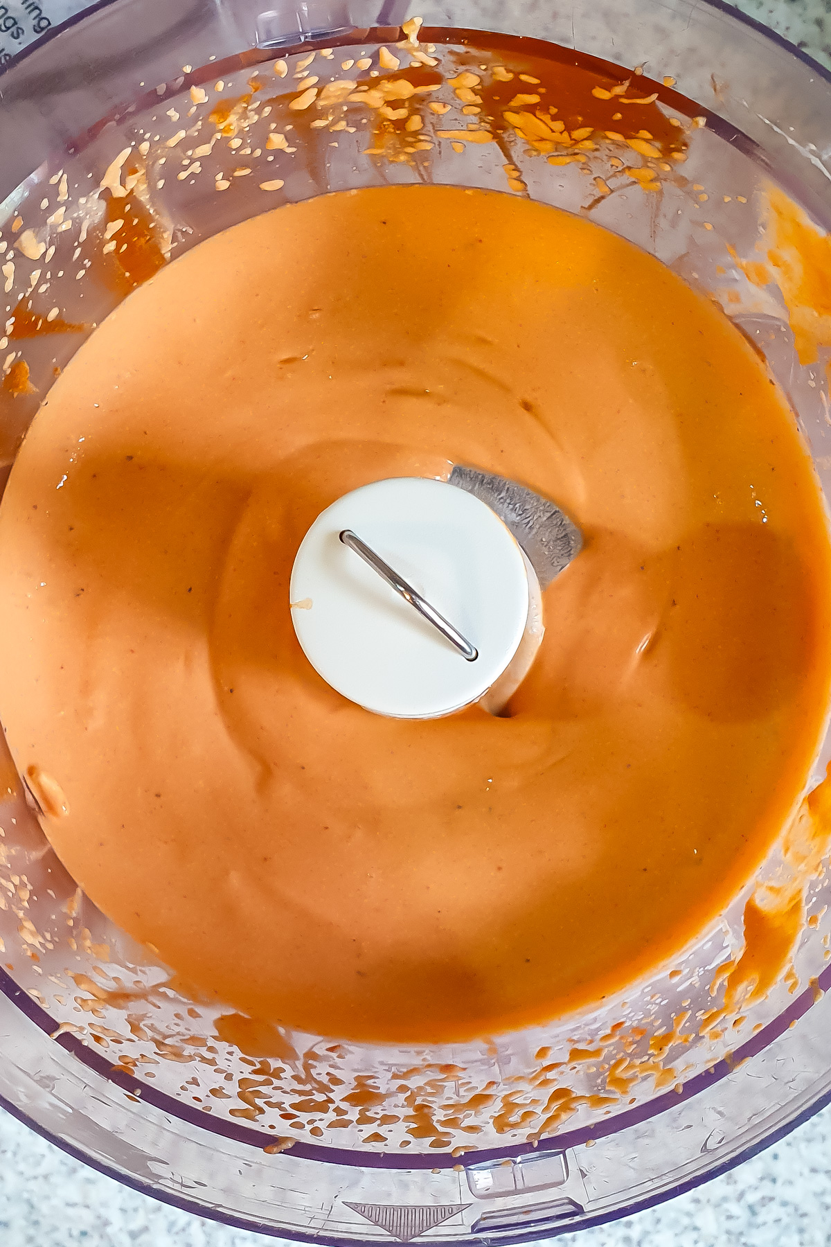 A food processor bowl filled with smooth, orange-colored Cottage cheese Buffalo Dip, with some residue on the sides and the blade visible in the center.