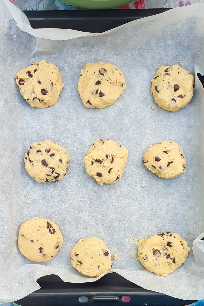 Nine unbaked Chocolate Chip Cookies are evenly spaced on a baking tray lined with parchment paper.