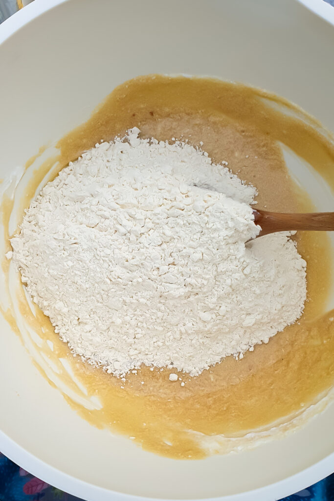 A mixing bowl with flour sitting on top of a wet cookie batter mixture, being stirred with a wooden spoon.
