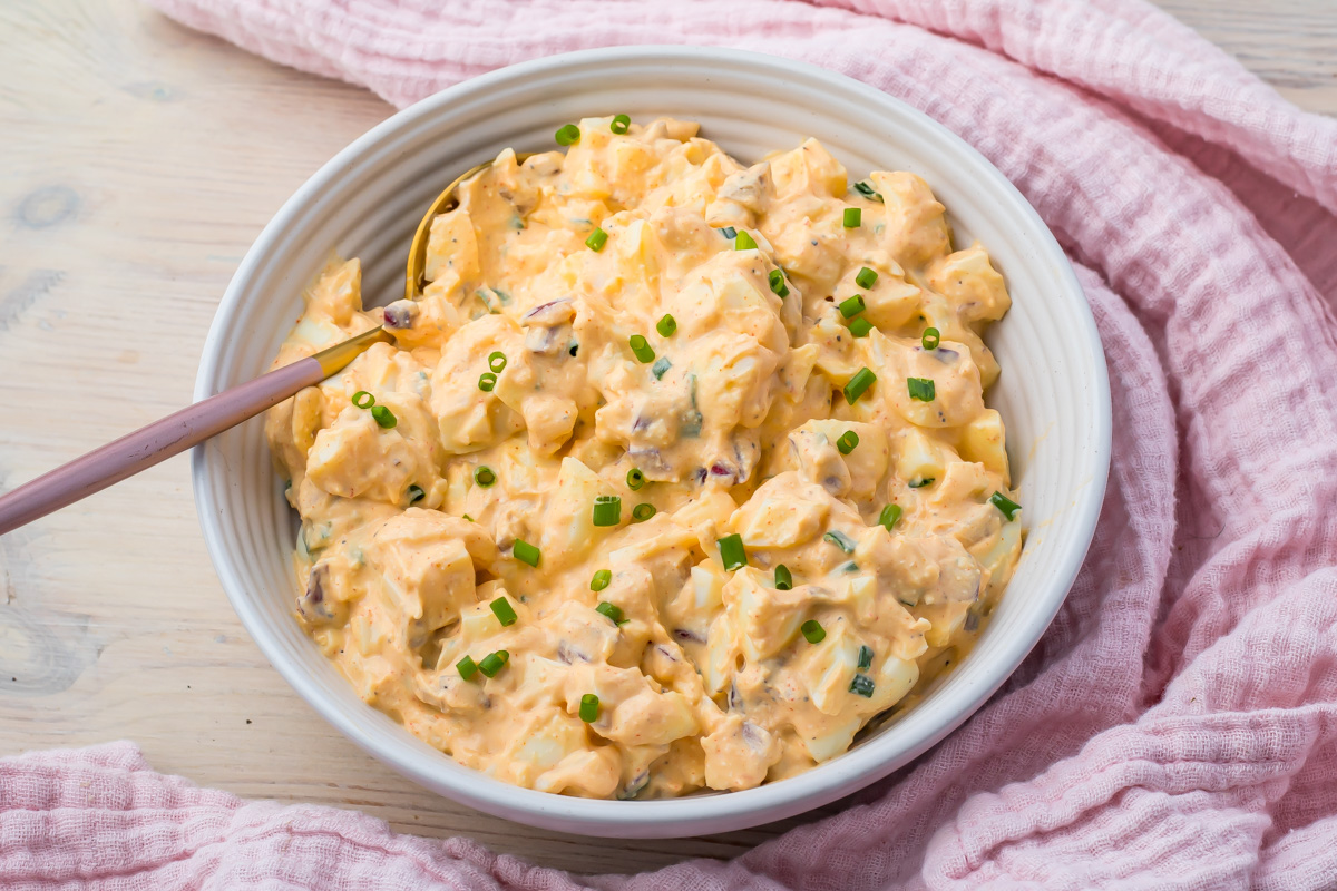 A bowl of cottage cheese egg salad with chopped chives on top, is placed next to a pink textured cloth.
