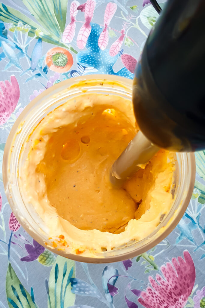 A hand blender whips up a creamy orange mixture—perfect for cottage cheese or your favorite egg salad recipe—set against a colorful, floral-patterned tablecloth.