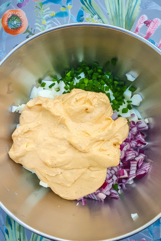 A metal bowl containing chopped green onions, chopped red onions, chopped egg whites, and a large dollop of creamy yellow sauce sits on a floral tablecloth—perfect for an easy egg salad recipe.