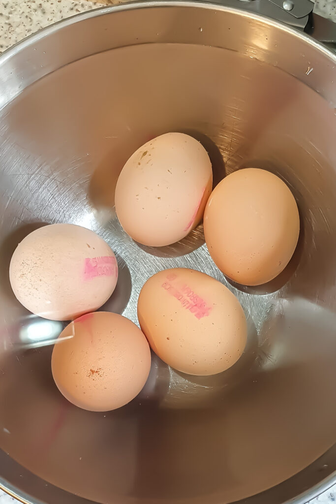 Five brown eggs with pink stamps are placed in a stainless steel mixing bowl, ready to be transformed into a delicious egg salad recipe.