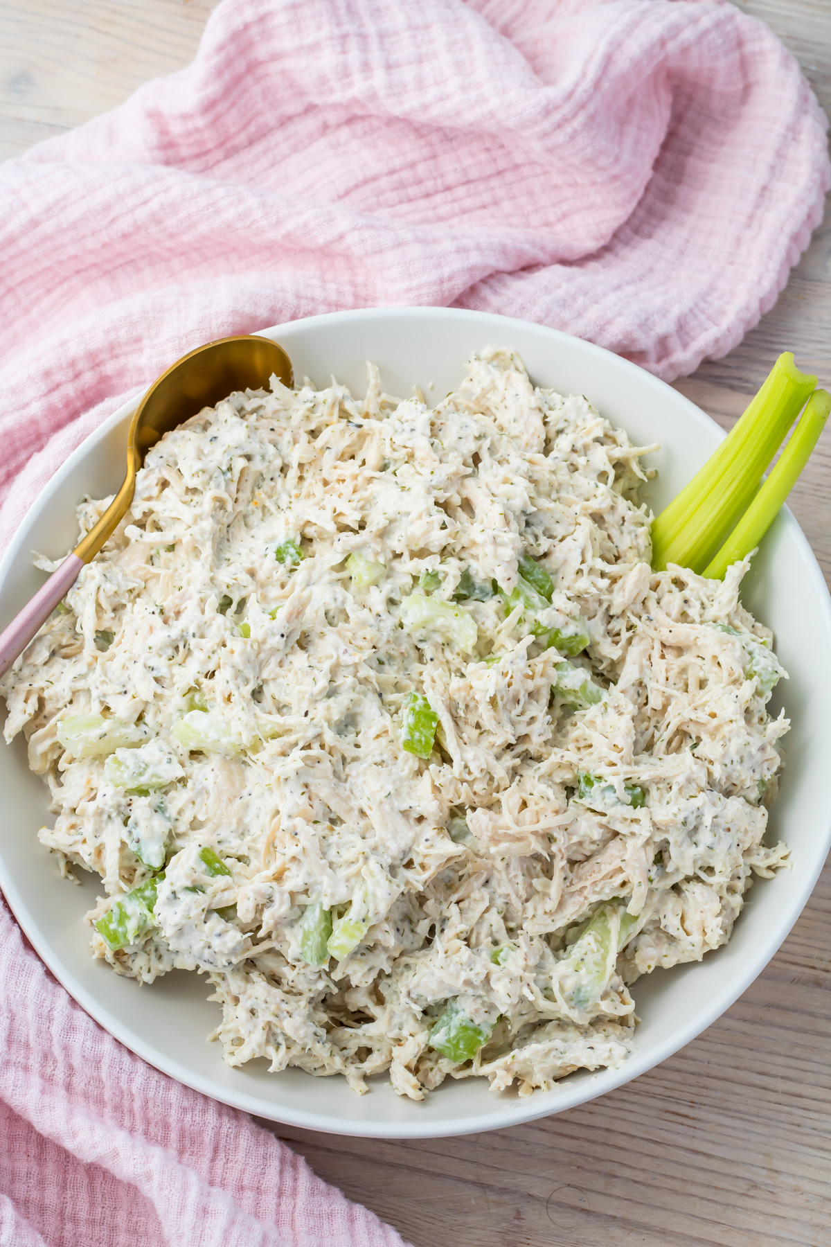 A white bowl filled with Ranch Chicken Salad mixed with herbs and celery, with celery sticks and a gold spoon on the side, placed on a light wooden surface with a pink cloth.