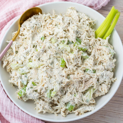 A white bowl filled with Ranch Chicken Salad, mixed with mayonnaise and chopped celery, sits on a light wooden surface with a pink cloth nearby.