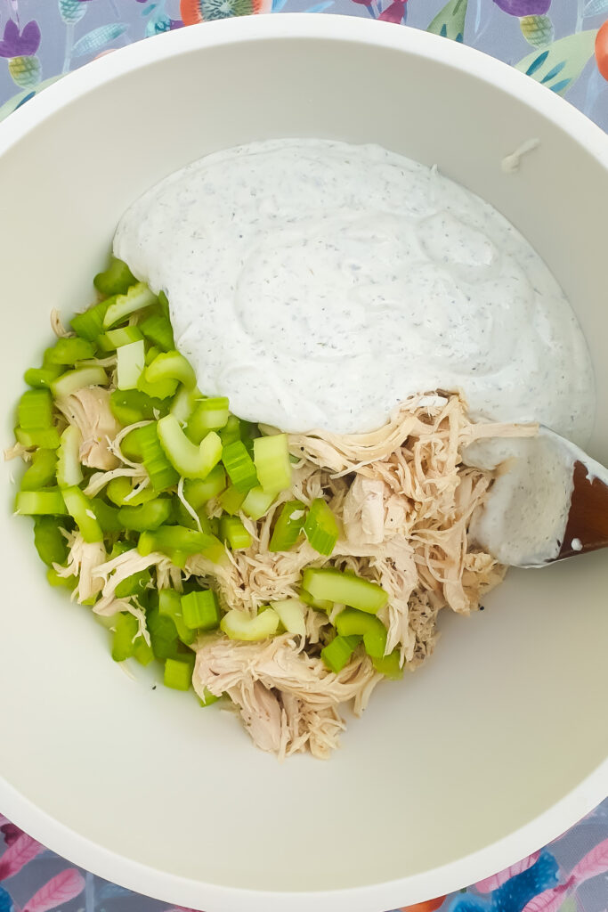 A white mixing bowl with chopped celery, shredded chicken, and creamy ranch dressing being combined with a spatula for a delicious Ranch Chicken Salad.