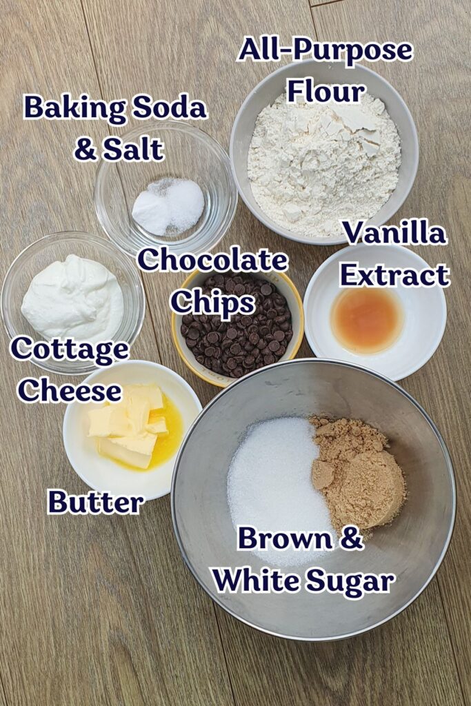 Top-down view of ingredients for cottage cheese chocolate chip cookies in labeled bowls: all-purpose flour, vanilla extract, brown & white sugar, butter, cottage cheese, chocolate chips, baking soda & salt.