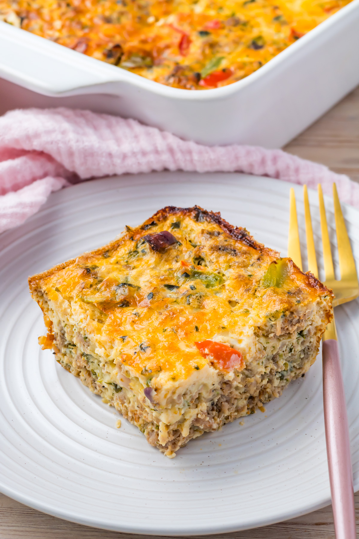 A slice of Cottage Cheese Egg Bake with vegetables and cheese on a white plate, next to a gold fork. A pink towel and casserole dish are in the background.