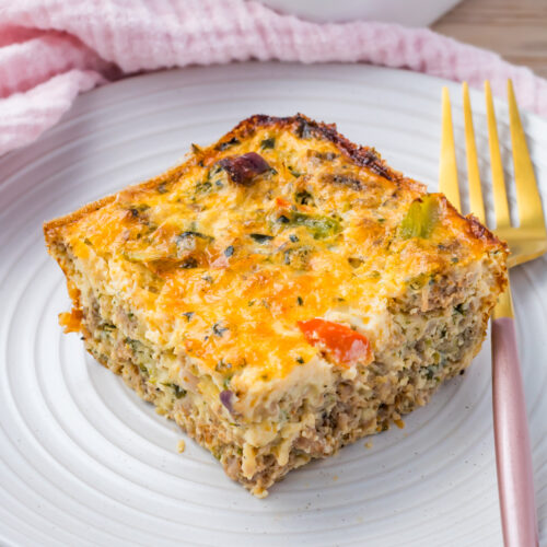 A square slice of cottage cheese egg bake with vegetables and cheese sits on a white plate next to a gold fork.