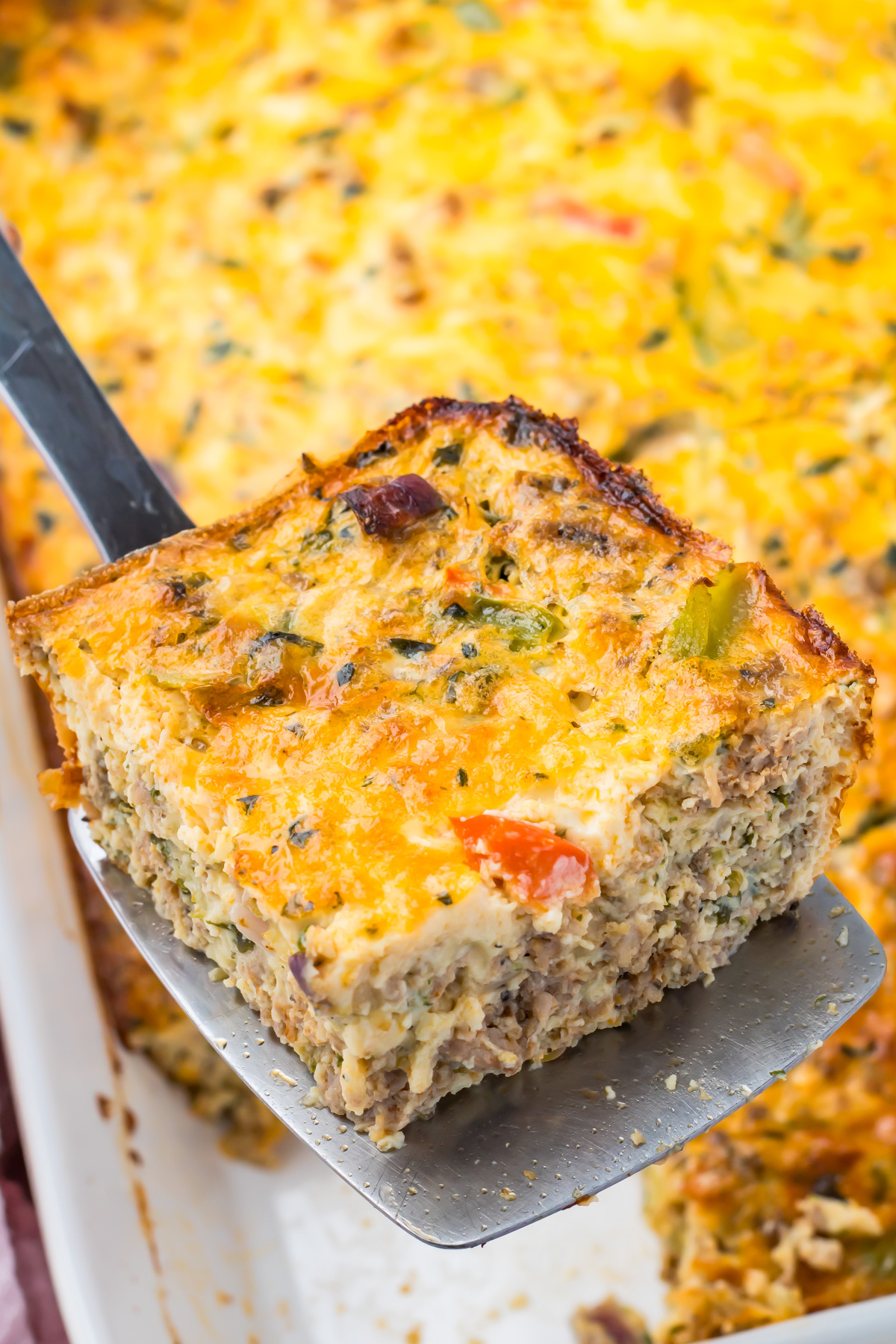 A close-up of a metal spatula lifting a square slice of egg bake breakfast casserole, featuring visible vegetables, cheese, and meat—plus creamy cottage cheese—from a white baking dish.